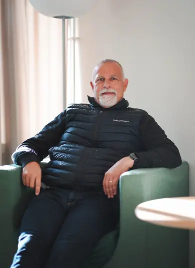 Man med svart väst som sitter i en fotölj och tittar in i kameran